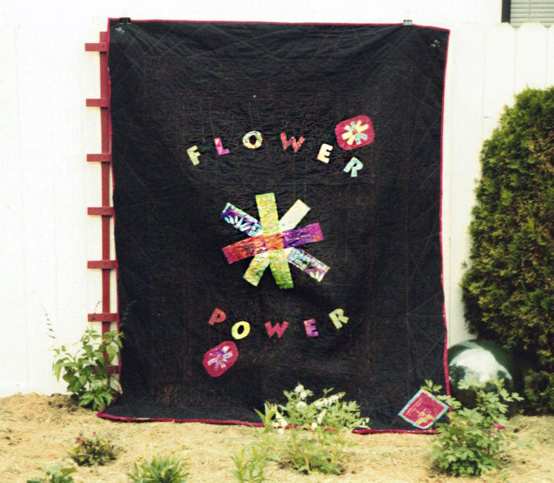 A quilt hanging on a red trellis outdoors. The quilt is a large black one with flower power and a large stargazey quilts daisiez design on it which is multicoloured