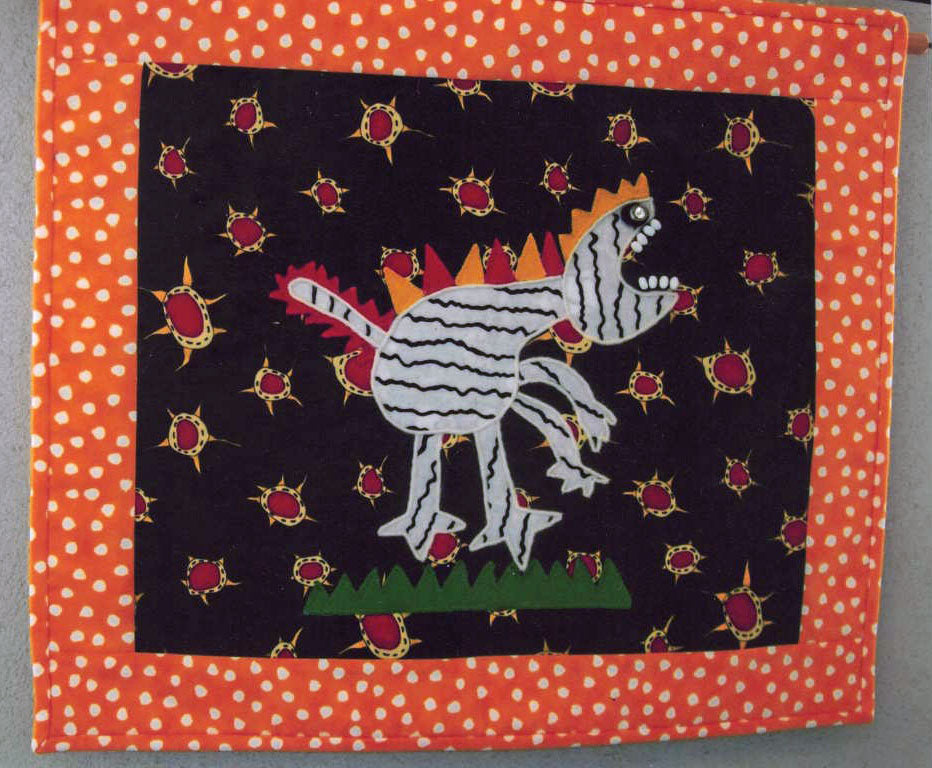 A small quilt with an orange dotted border. The centre block is a picture of an abstract dinosaur with spikes and white bobbles for teeth