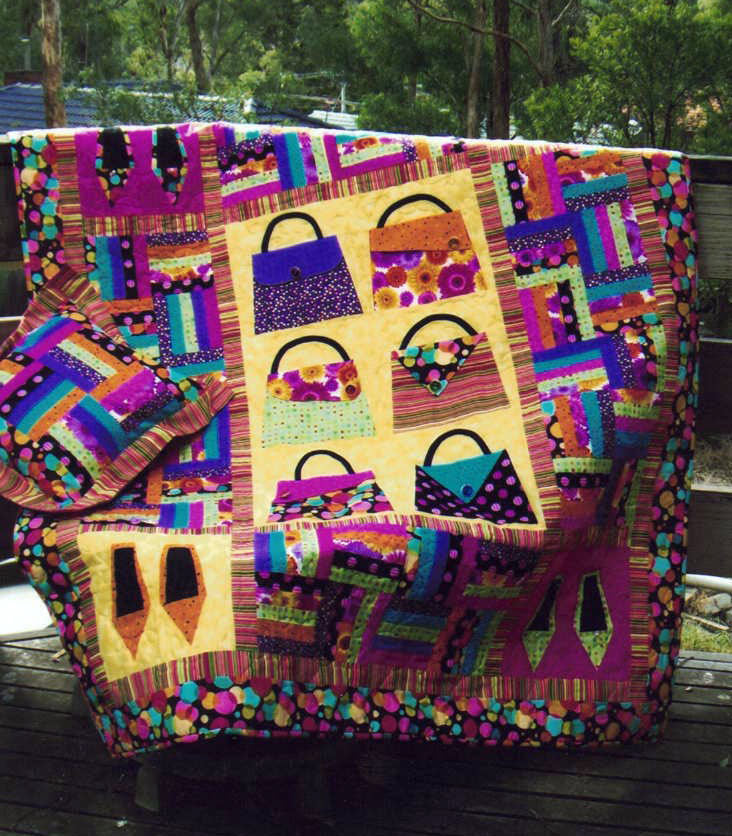 A bright pink and yellow large quilt hanging on a clothes line outdoors. There are handbags and shoes sewn into the quilt