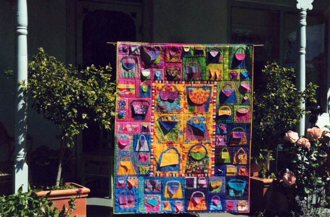 A photograph of a large quilt hanging on the front porch of a house. The quilt is colourful with lots of different sized handbags with flaps as the handbag openings