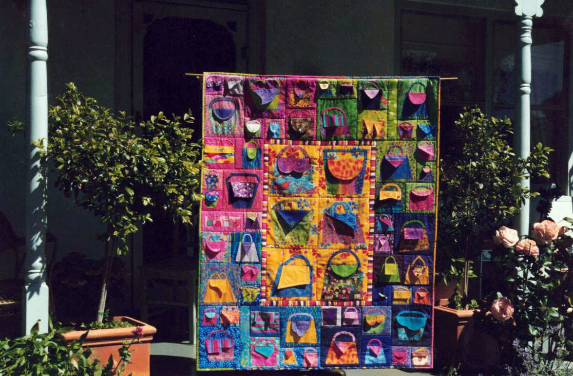 A photograph of a large quilt hanging on the front porch of a house. The quilt is colourful with lots of different sized handbags with flaps as the handbag openings