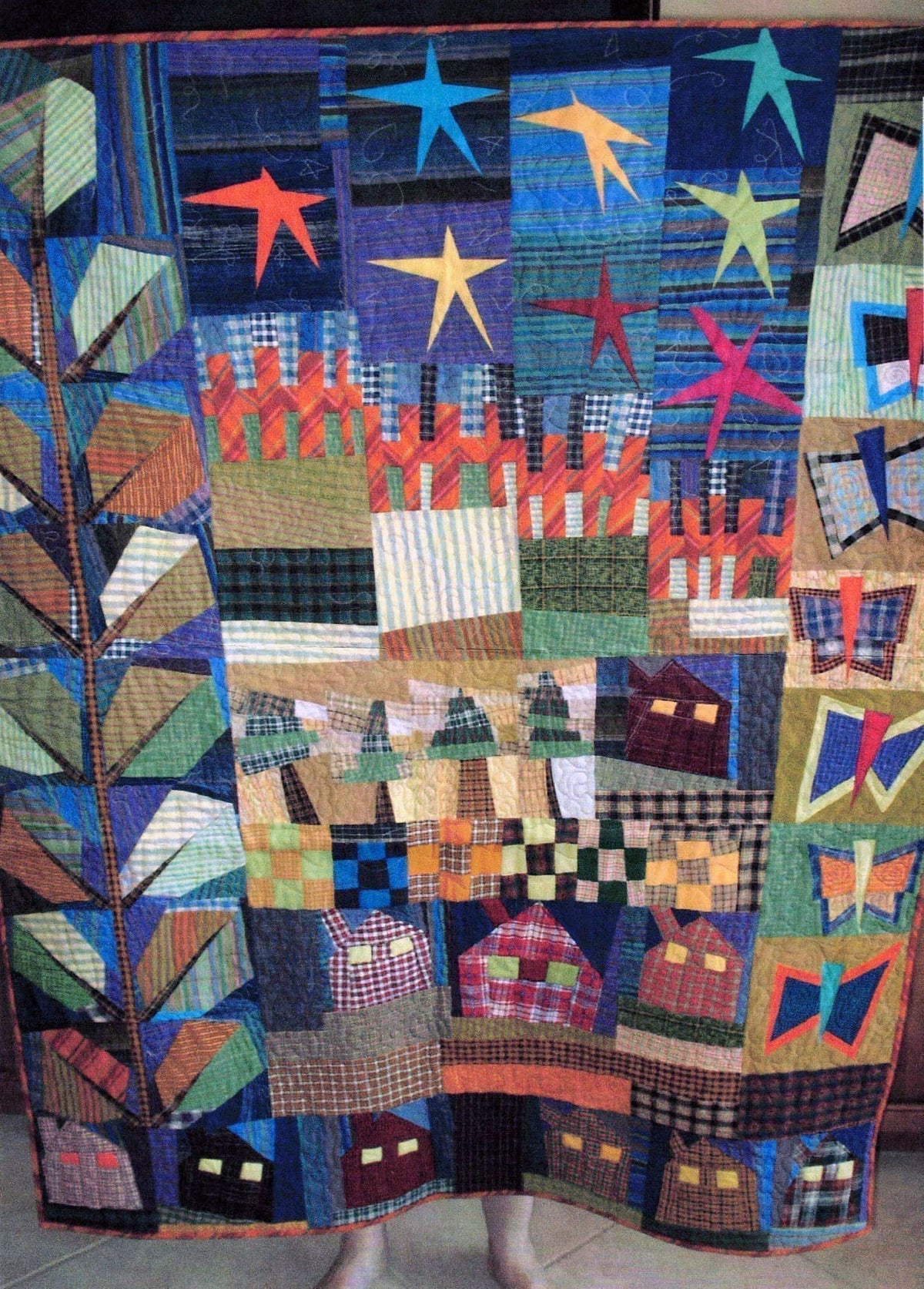 A photo of a home-made quilt in many colours. A sky, stars, fences, houses, leaves and butterflies.