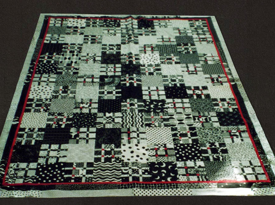 A photograph of a quilt laid on on a carpeted floor. The quilt is black and white grids like a tic tac toe grid. Ther fabrics are black and white with red splashes of colour